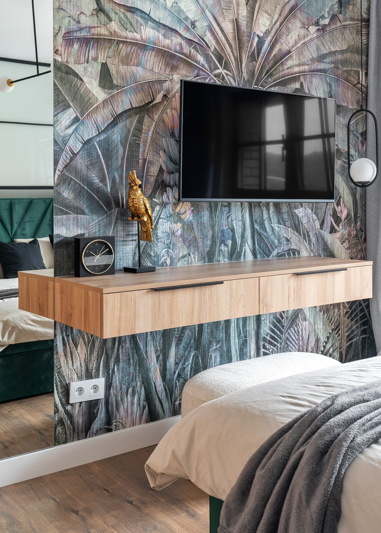 Nowoczesna sypialnia z tapetą w motywy tropikalne, podwieszana szafka RTV z drewnianym dekorem, złota figurka papugi i czarny zegar na blacie, odbicie łóżka w lustrze.