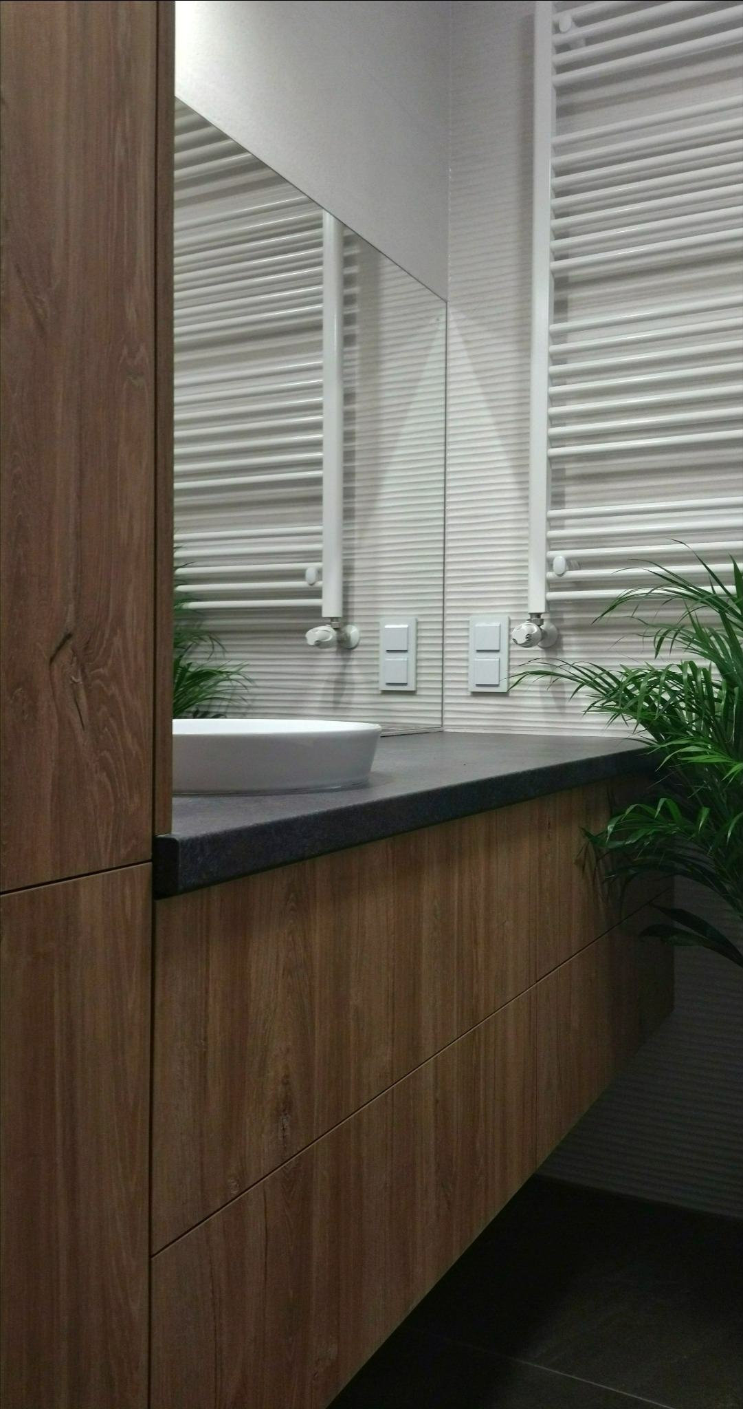 Nowoczesna łazienka z drewnianą szafką pod umywalką, ciemnym blatem, białym grzejnikiem drabinkowym i rośliną doniczkową w tle.