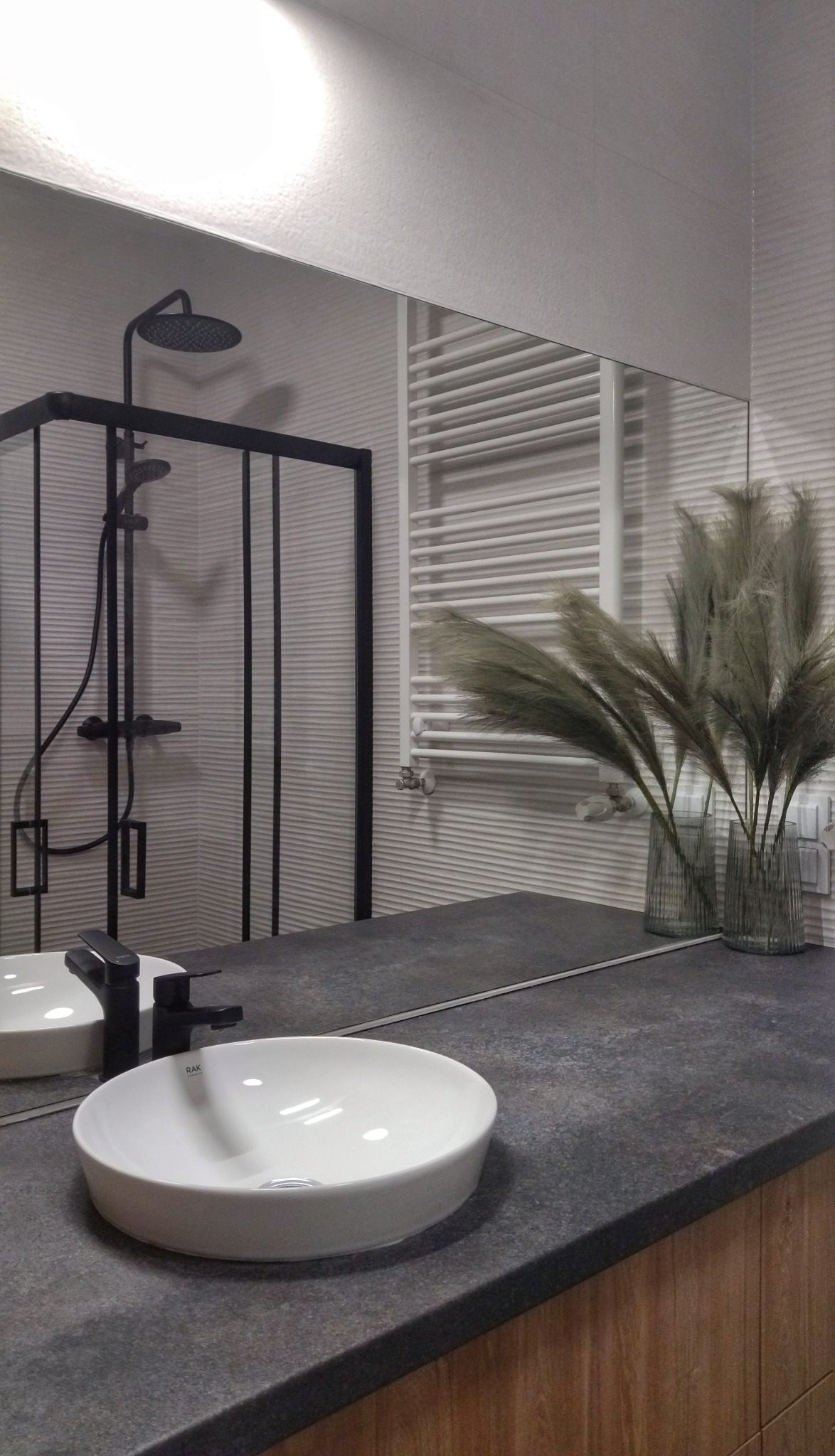 Nowoczesna łazienka z okrągłą umywalką nablatową, czarną armaturą i kabiną prysznicową z czarnymi ramami, odbijająca się w lustrze, z dekoracyjnymi trawami w wazonach.