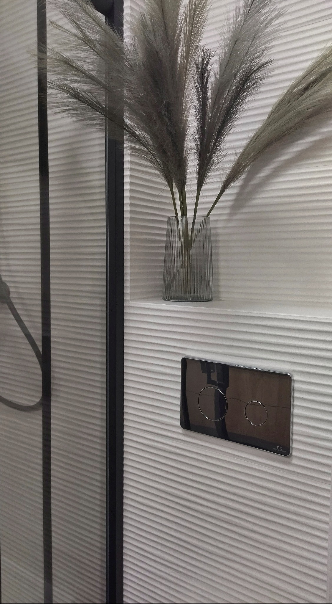 Nowoczesna łazienka z teksturalną białą ścianą, szklanym prysznicem z czarną ramą, ozdobnym wazonem z trawami pampasowymi i chromowanym przyciskiem spłuczki.