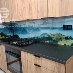 Nowoczesna kuchnia z drewnopodobnymi szafkami, czarnym blatem i szklanym panelem ściennym z motywem górskiego krajobrazu, widoczna płyta gazowa i piekarnik.