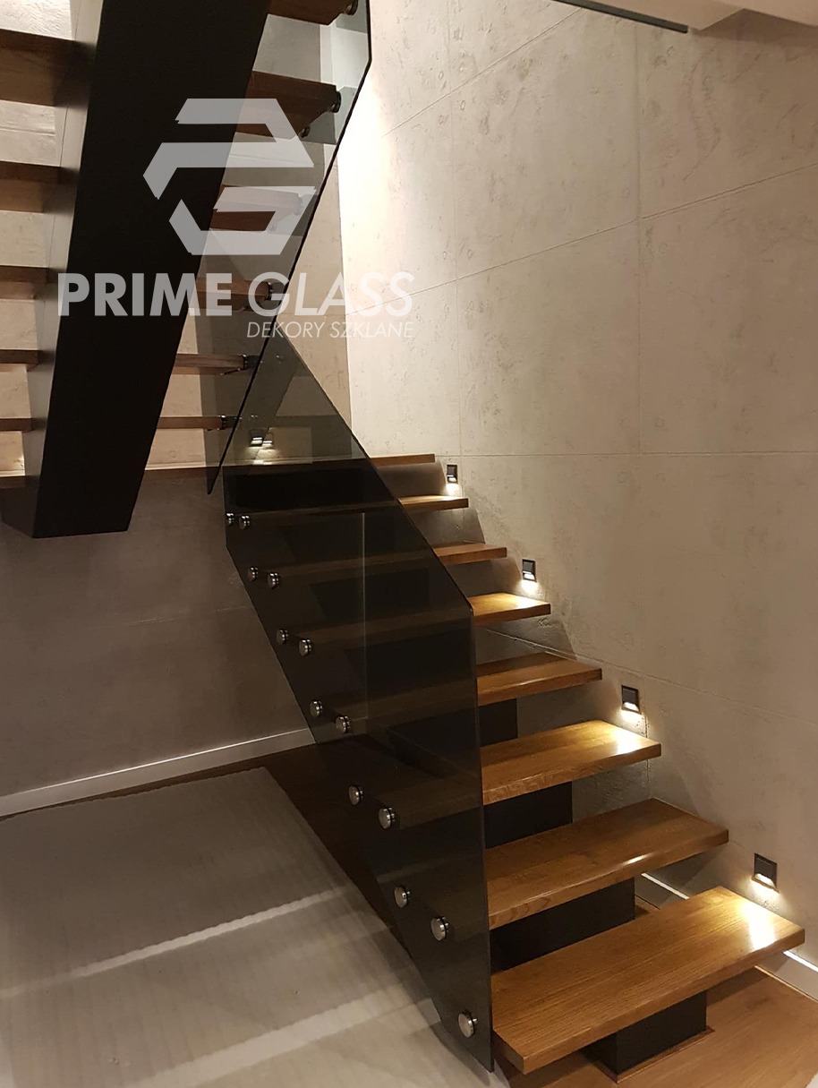 Nowoczesne schody z drewnianymi stopniami i szklaną balustradą mocowaną punktowo, oświetlone małymi ledami w ścianie, z widocznym logo firmy szklarskiej na górze.