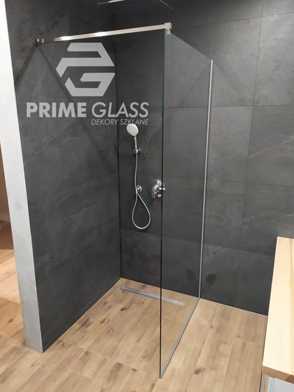 Nowoczesna kabina prysznicowa ze szkła, z logo firmy Prime Glass na ciemnych, marmurkowych płytkach, drewnopodobna podłoga, odpływ liniowy.