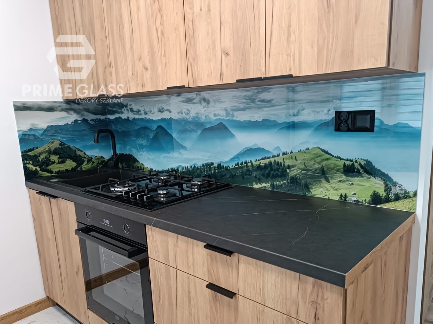 Nowoczesna kuchnia z drewnopodobnymi szafkami, czarnym blatem i szklanym panelem ściennym z motywem górskiego krajobrazu, widoczna płyta gazowa i piekarnik.