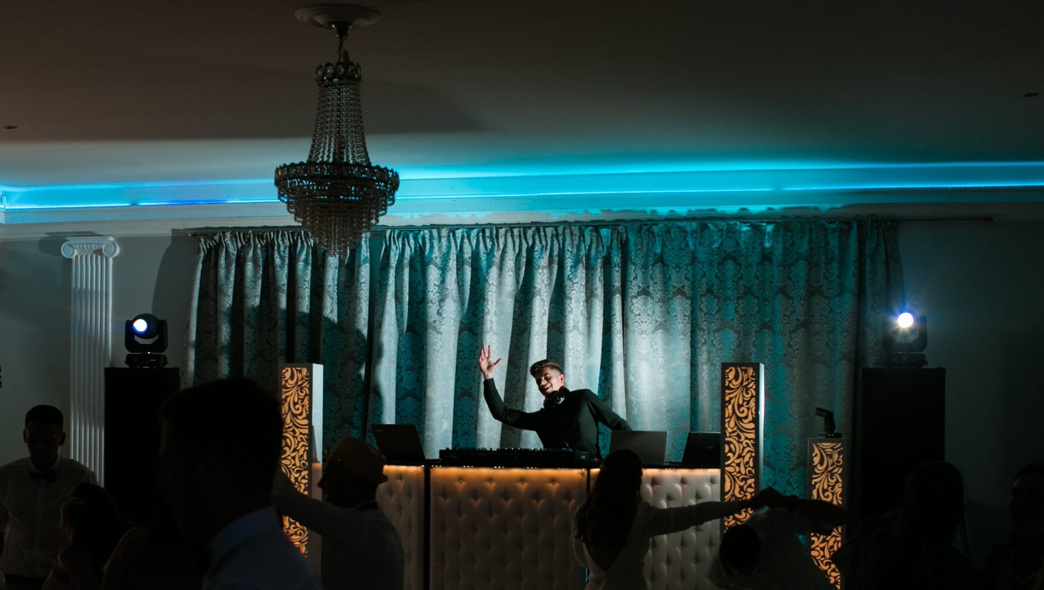 DJ w czarnej koszuli, z podniesioną ręką, za konsolą DJ-ską oświetloną od dołu, w tle dekoracyjne kolumny, żyrandol i tańczący tłum na sali balowej z niebieskim oświetleniem.