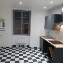 Nowoczesna kuchnia z czarno-białą podłogą w szachownicę, szarymi szafkami i drewnianym blatem, widok na okno z szarymi ramami i biały grzejnik pod nim.