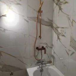 Bateria wannowa w kolorze miedzi z deszczownicą na ścianie wyłożonej płytkami imitującymi marmur, widoczne fragmenty wanny.