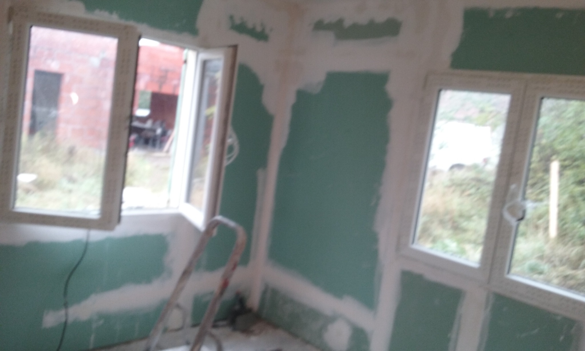 Wnętrze pokoju z częściowo położonymi płytami gipsowo-kartonowymi, widoczne szpachlowane łączenia, aluminiowa drabina oparta o ścianę, dwa okna PCV z widokiem na zewnątrz.