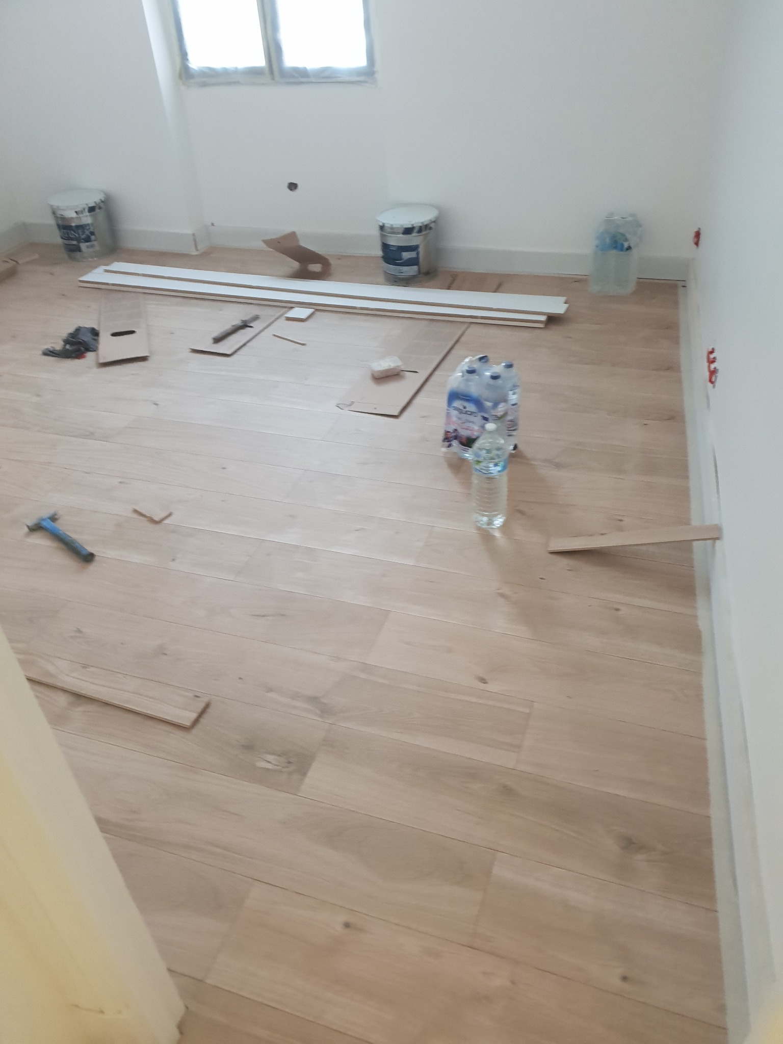 Montaż jasnej, drewnopodobnej podłogi w pustym pomieszczeniu, widoczne narzędzia, listwy przypodłogowe, farby i woda mineralna.