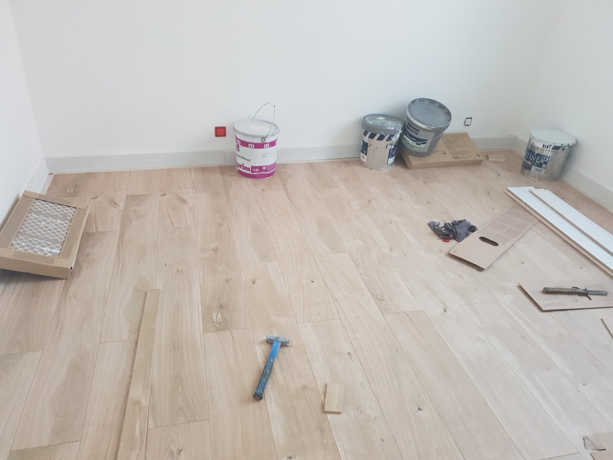 Podłoga z jasnego drewna w trakcie układania, widoczne narzędzia, puszki z farbą i listwy przypodłogowe.