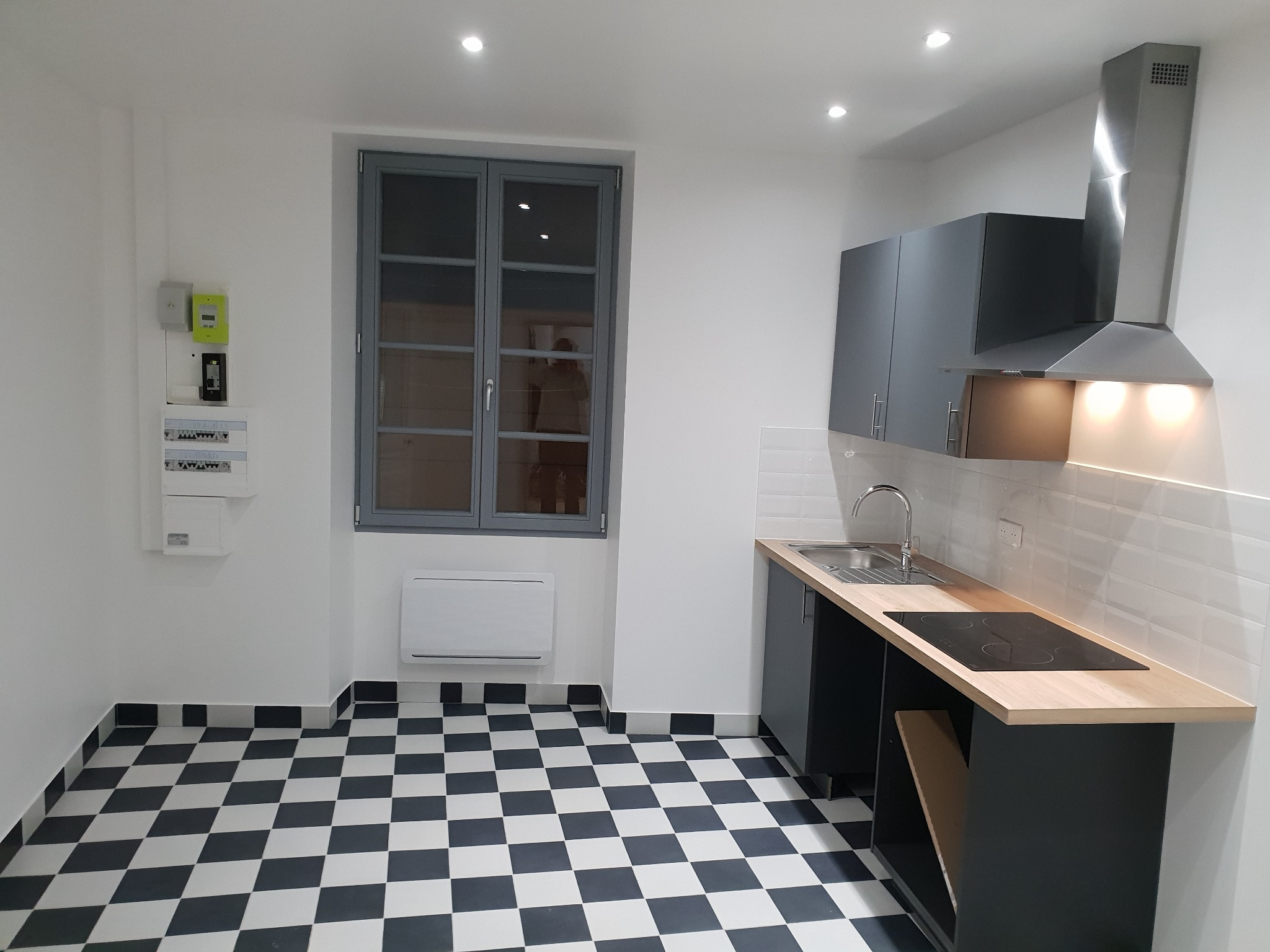 Nowoczesna kuchnia z czarno-białą podłogą w szachownicę, szarymi szafkami i drewnianym blatem, widok na okno z szarymi ramami i biały grzejnik pod nim.