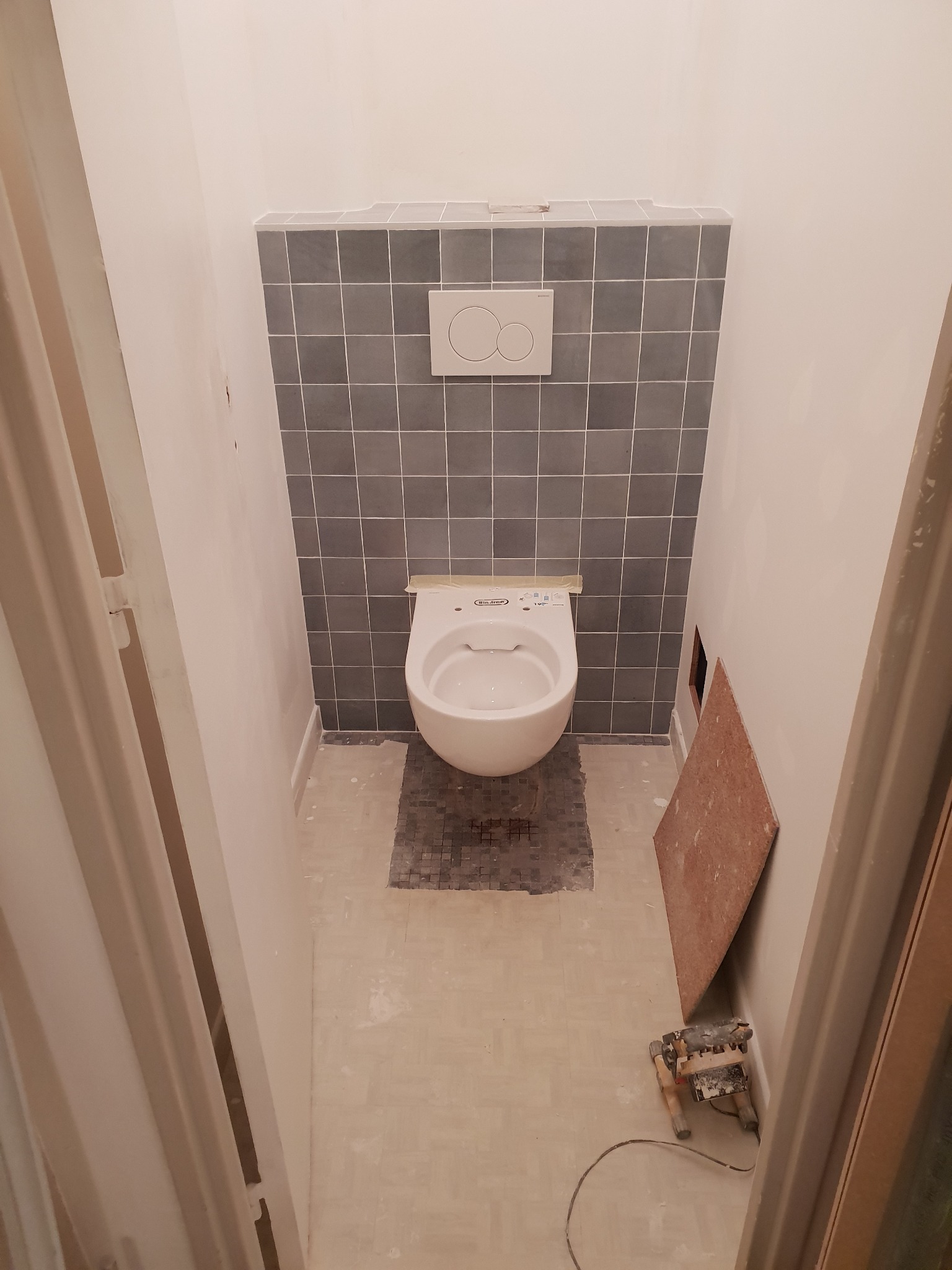 Wisząca toaleta z szarą mozaiką pod spodem, szare płytki na ścianie za toaletą, widoczne narzędzia i płyta kartonowo-gipsowa, ściany przygotowane pod malowanie.