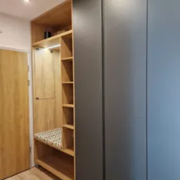 Wnęka z siedziskiem i półkami z jasnego drewna, wkomponowana w szarą zabudowę szafy w przedpokoju, obok drzwi z drewnianą okleiną.