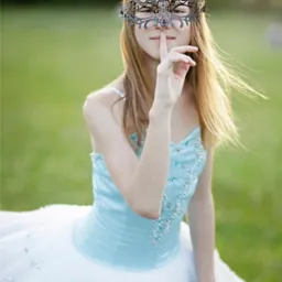 Młoda kobieta w baletowej sukience i ozdobnej masce na tle zielonej łąki, z palcem przy ustach w geście uciszenia.