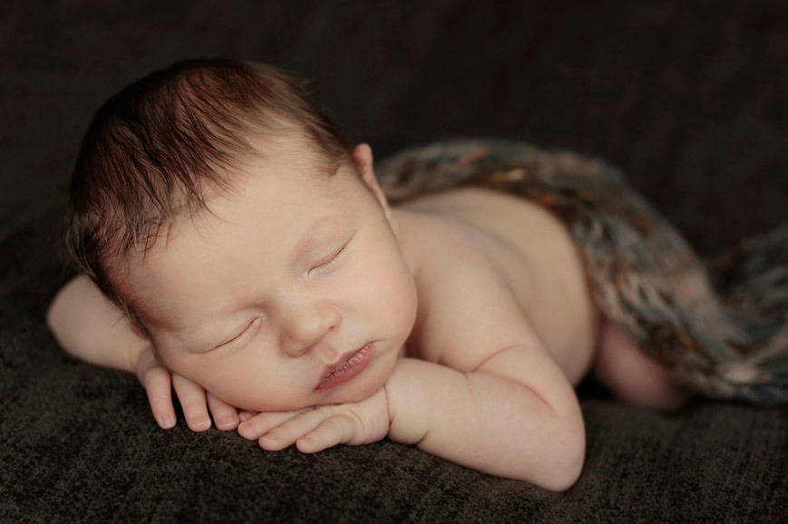 Śpiący noworodek z ciemnymi włosami, ułożony na dłoniach na ciemnobrązowym tle, przykryty wzorzystym kocykiem.