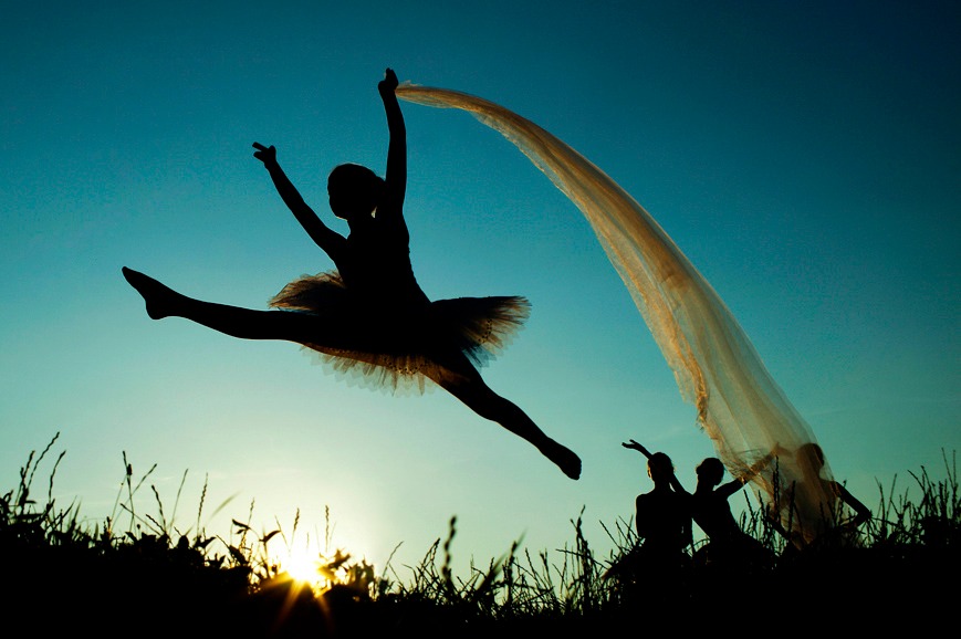 Sylwetka baletnicy w tutu wykonującej skok w rozkroku na tle błękitnego nieba i zachodzącego słońca, z powiewającą tkaniną i grupą osób w dole kadru.