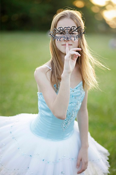 Młoda kobieta w baletowej sukience i ozdobnej masce na tle zielonej łąki, z palcem przy ustach w geście uciszenia.
