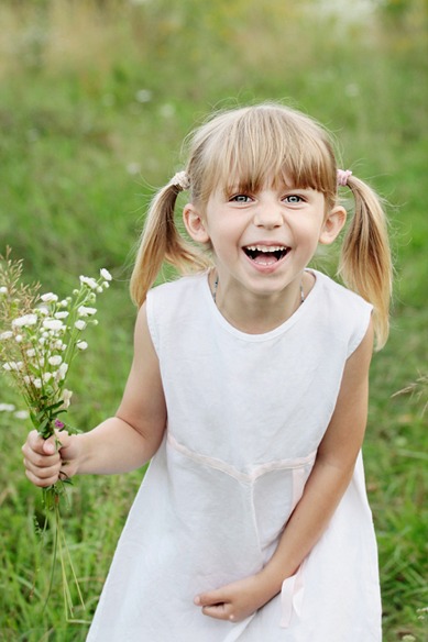 Uśmiechnięta dziewczynka w białej sukience trzyma bukiet polnych kwiatów na tle zielonej łąki.
