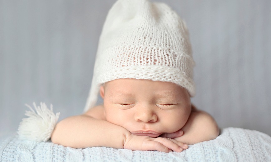 Śpiący noworodek w białej, dzierganej czapeczce z pomponem, leżący na błękitnym, dzierganym kocyku, z zamkniętymi oczami i rączkami pod policzkami.