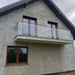 Dom w stanie surowym z balkonem zabezpieczonym szklaną balustradą z elementami ze stali nierdzewnej i brązowymi ramami okiennymi.