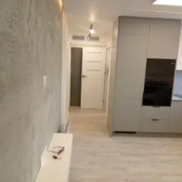 Szara ściana z efektem dekoracyjnym, jasna podłoga imitująca drewno, nowoczesna zabudowa kuchenna w odcieniach szarości z czarnym piekarnikiem i białe drzwi w tle.