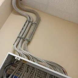 Szereg szarych kabli prowadzonych w metalowych korytkach, wychodzących z otworu w suficie podwieszanym i wchodzących do otwartej skrzynki rozdzielczej na ścianie, widok z dołu.