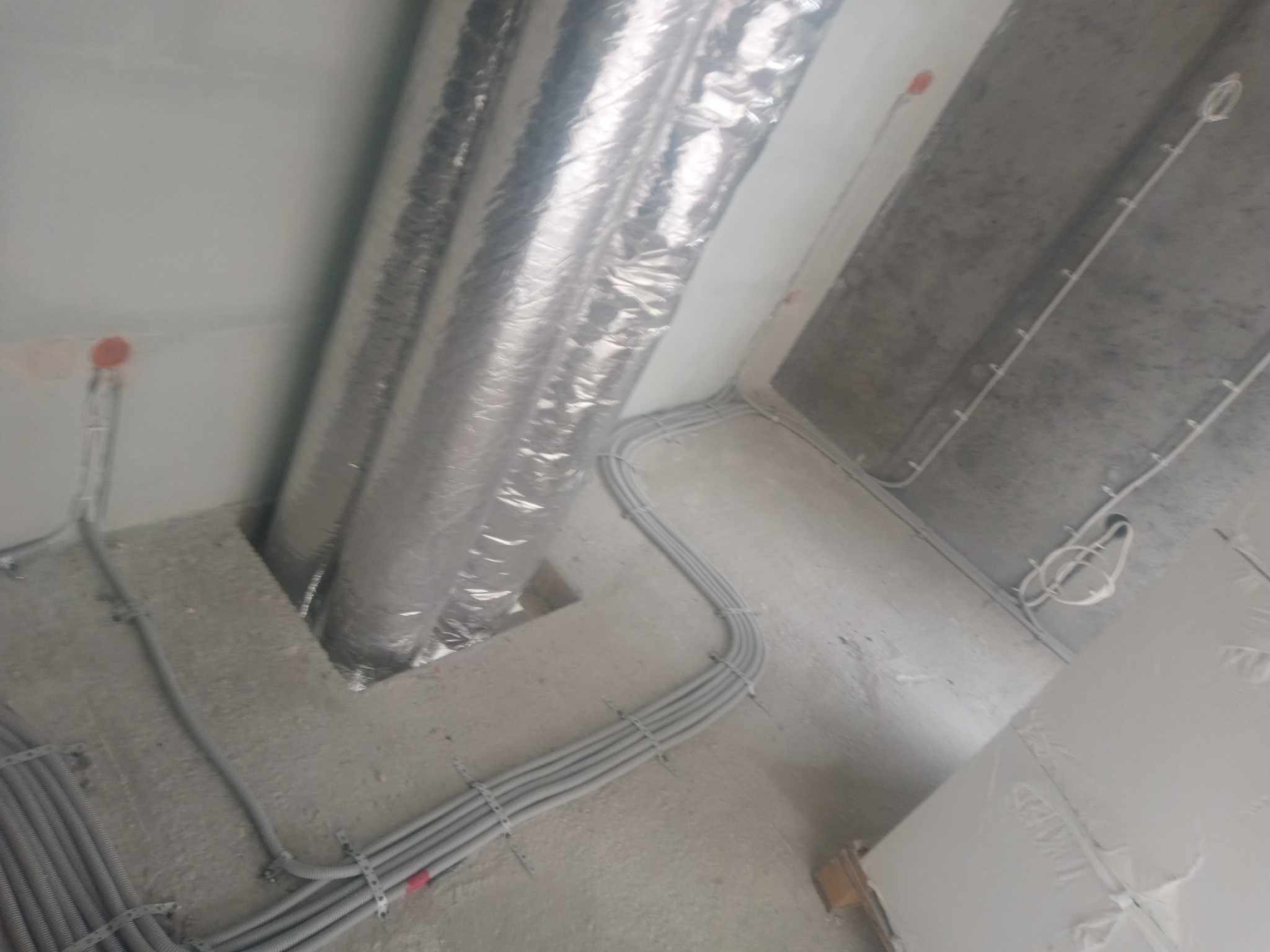 Surowe wnętrze w trakcie prac: widoczne rury wentylacyjne w srebrnej izolacji, szare peszle z przewodami elektrycznymi przymocowane do surowego sufitu i ścian, czerwone puszki instalacyjne.