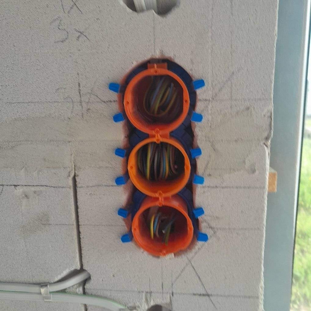 Potrójna puszka elektryczna z przewodami w ścianie z betonu komórkowego, zabezpieczona niebieskimi kołkami, widoczne oznaczenia ołówkiem na ścianie.