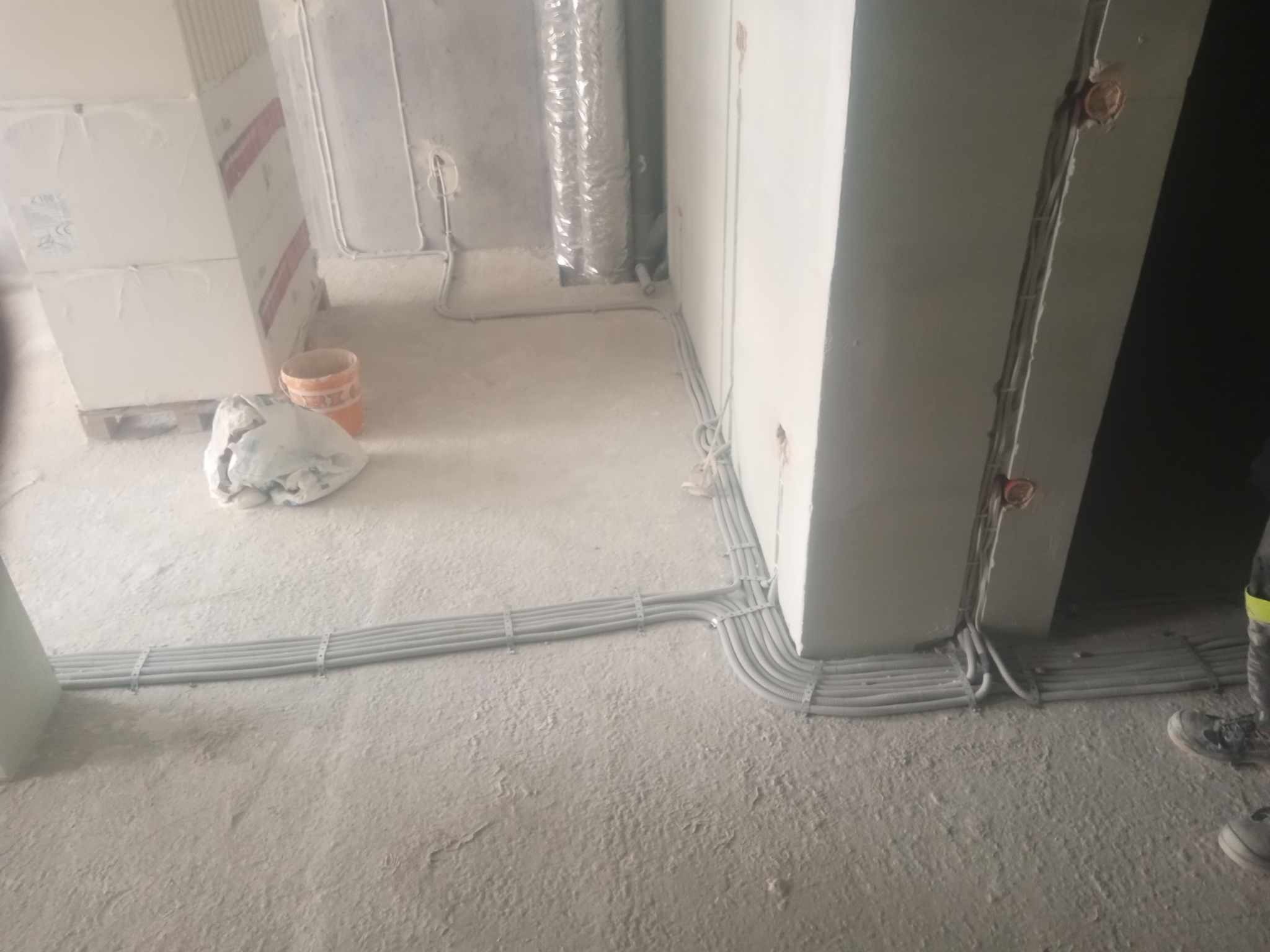 Szare przewody elektryczne ułożone wzdłuż podłogi w surowym wnętrzu, częściowo przymocowane uchwytami, prowadzone wzdłuż ściany, widoczny fragment instalacji wentylacyjnej i materiały budowlane.