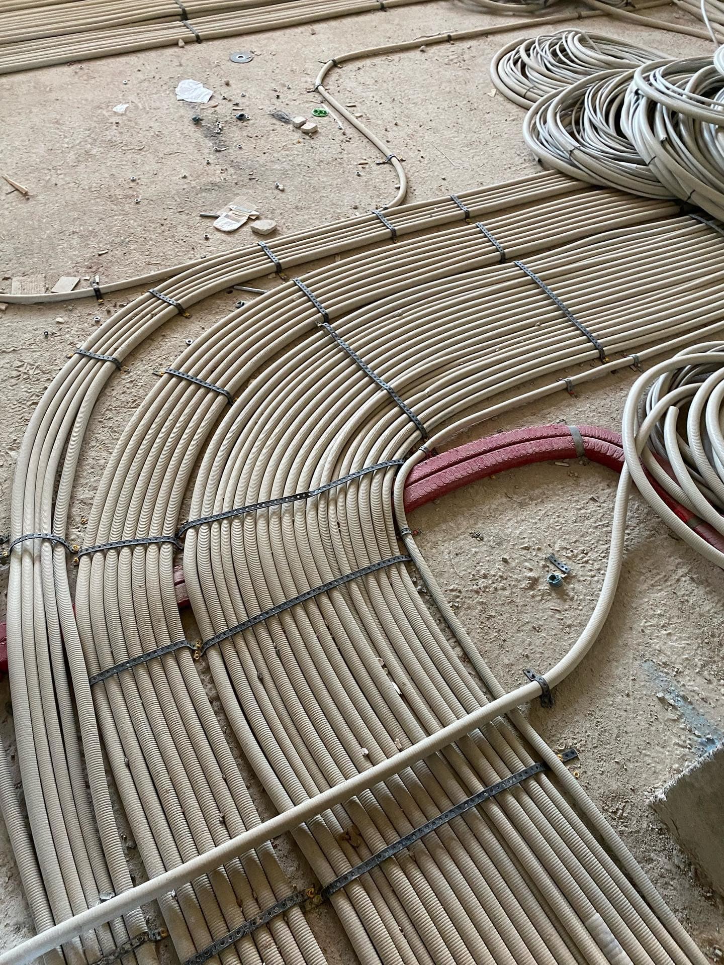 Rury karbowane ułożone równolegle na szorstkiej, betonowej podłodze budowy, spięte czarnymi opaskami, z widocznymi fragmentami rur w kolorze czerwonym.