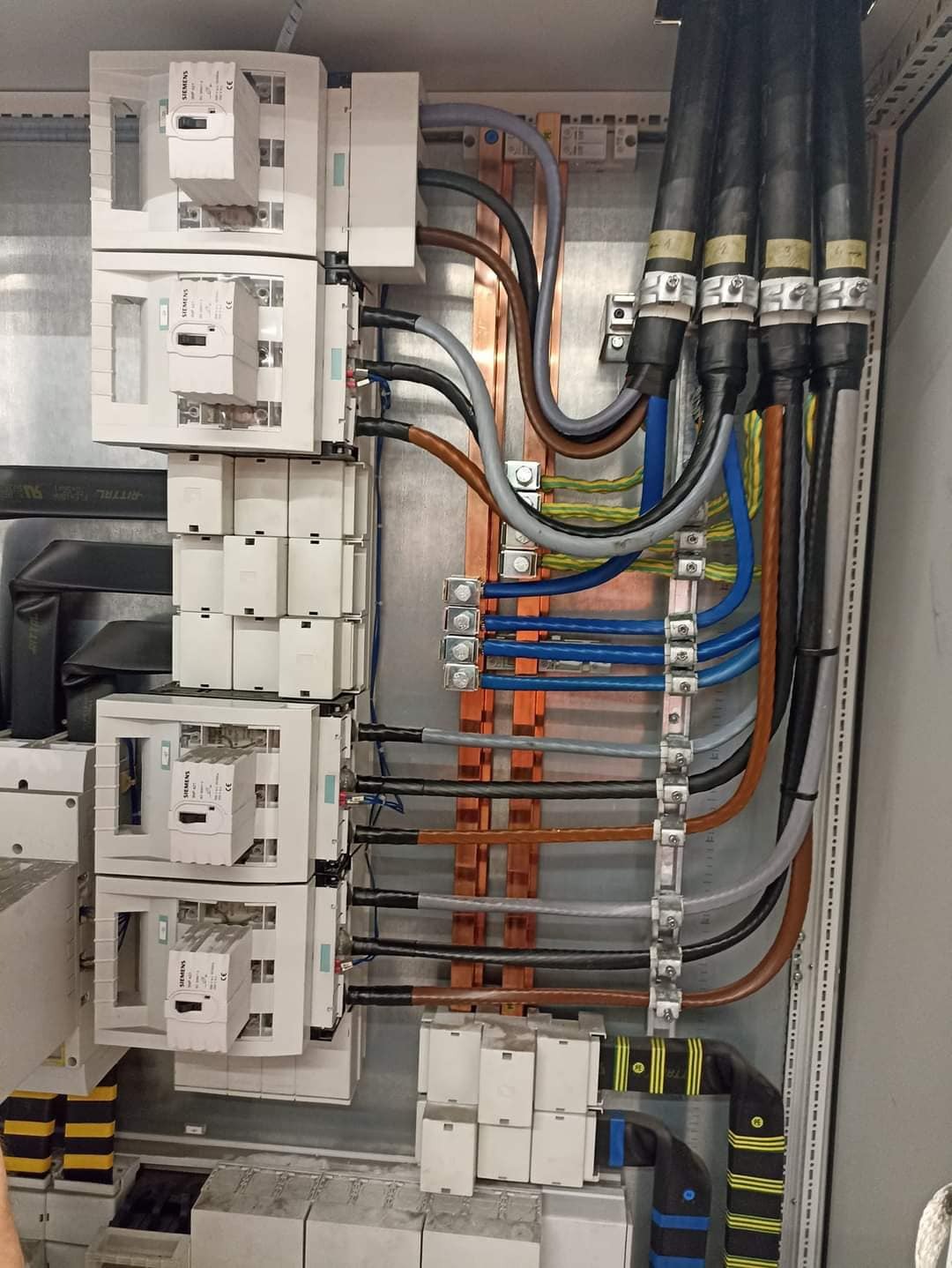 Rozdzielnica elektryczna z zainstalowanymi bezpiecznikami i okablowaniem, widoczne miedziane szyny i przewody w różnych kolorach, zabezpieczone peszlami z żółtymi paskami.