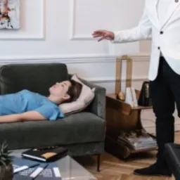 Kobieta leży na zielonej sofie podczas sesji hipnoterapii, mężczyzna w białej marynarce gestykuluje dłonią nad nią.