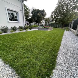 LuxGarden - Starannie utrzymany trawnik z równo przystrzyżoną trawą, obok ścieżka wysypana białym żwirem, widoczne elementy architektury ogrodowej i fragment domu.