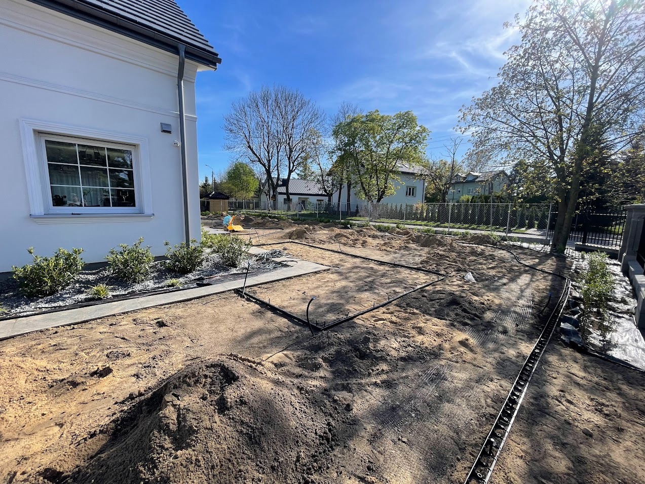 Przygotowanie terenu pod budowę: wykopana ziemia, piasek, wytyczone fundamenty, rury, obrzeża, widok z poziomu gruntu, dom z białym oknem w tle, błękitne niebo.