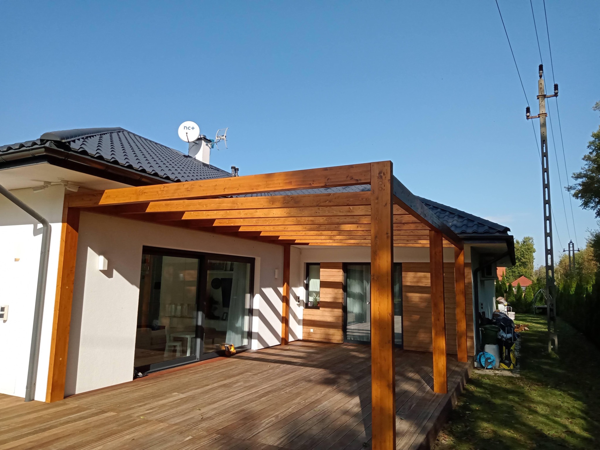 Drewniana pergola zadaszająca taras przylegający do nowoczesnego domu z białymi ścianami i grafitowym dachem, oświetlona popołudniowym słońcem, z widocznymi elementami wyposażenia ogrodu w tle.