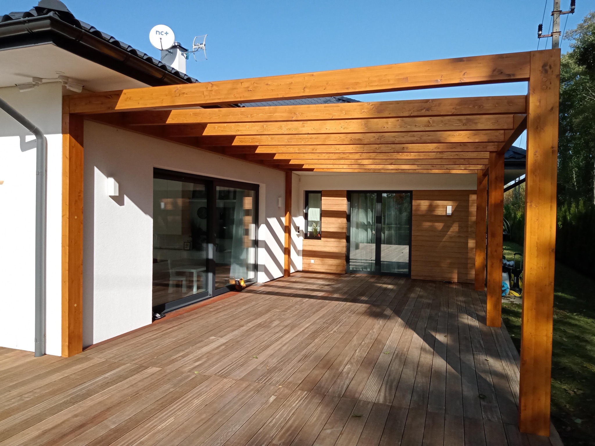 Nowoczesna drewniana pergola z zadaszeniem lamelowym, przylegająca do budynku z dużymi przesuwnymi oknami i drewnianym tarasem, oświetlona porannym słońcem.