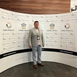 Mężczyzna w szarej marynarce pozuje na tle ściany z logotypami firmowymi 'Nasz Prąd S.A.' i 'Nasze Ciepło'.