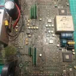 Zdemontowana, brudna płyta główna urządzenia elektronicznego z widocznymi elementami elektronicznymi, kondensatorami, układami scalonymi i złączami, leżąca na zielonej macie warsztatowej.