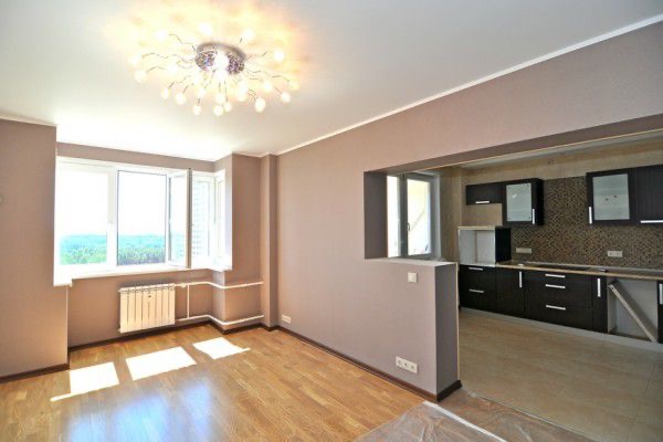 Jasne wnętrze po remoncie: parkiet, ściany w odcieniach beżu, nowoczesna lampa sufitowa, widok na kuchnię przez otwór w ścianie, okno z widokiem na zieleń.