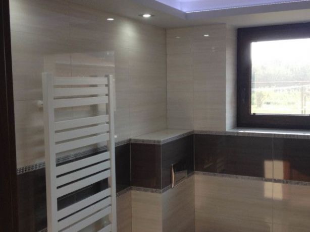 Wykończona łazienka z beżowo-brązowymi płytkami, białym grzejnikiem drabinkowym i oknem z ciemną ramą.