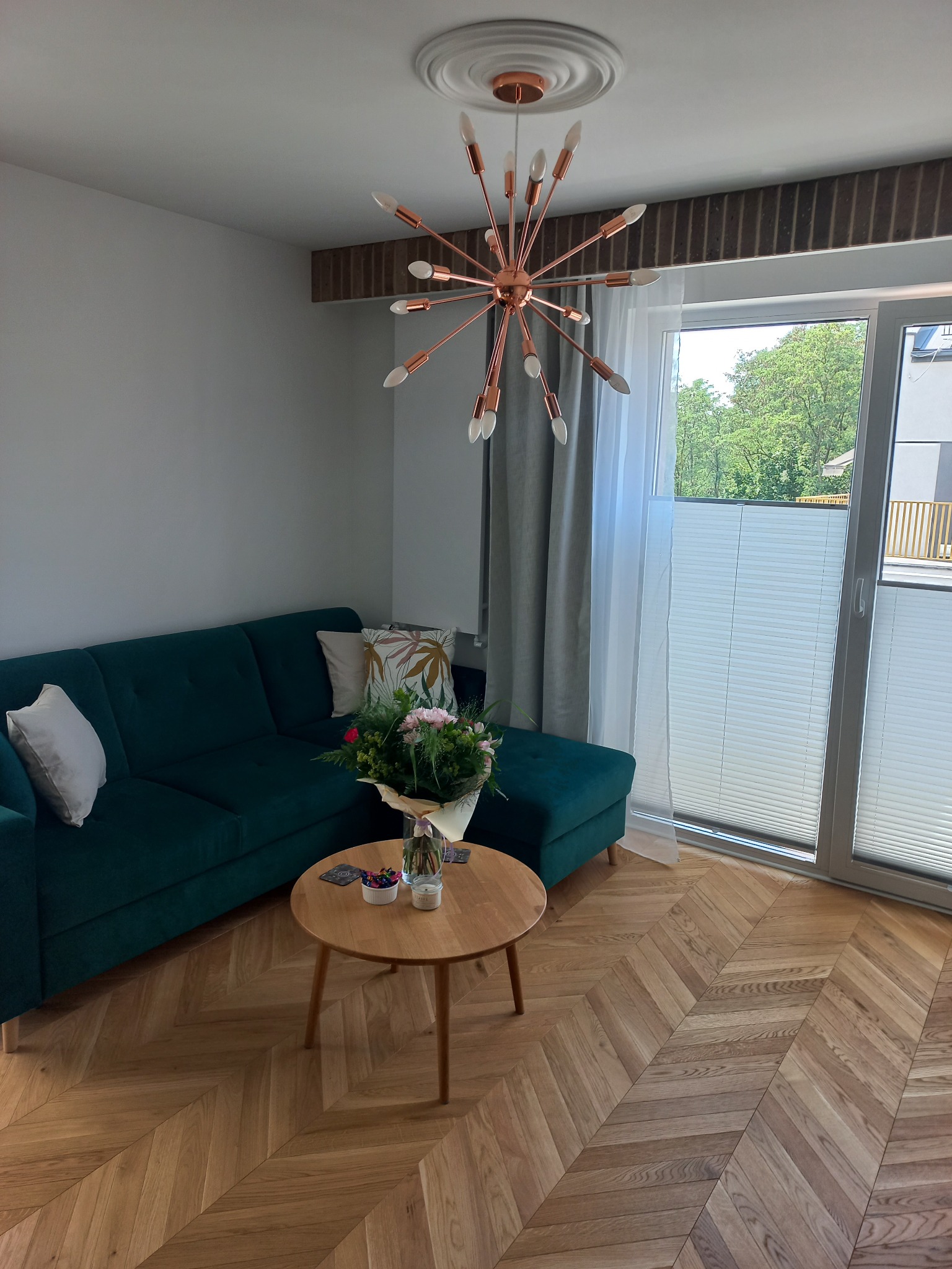 Salon z parkietem ułożonym w jodełkę, zieloną sofą i lampą w stylu retro, oświetlony naturalnym światłem z okna z plisowanymi roletami.