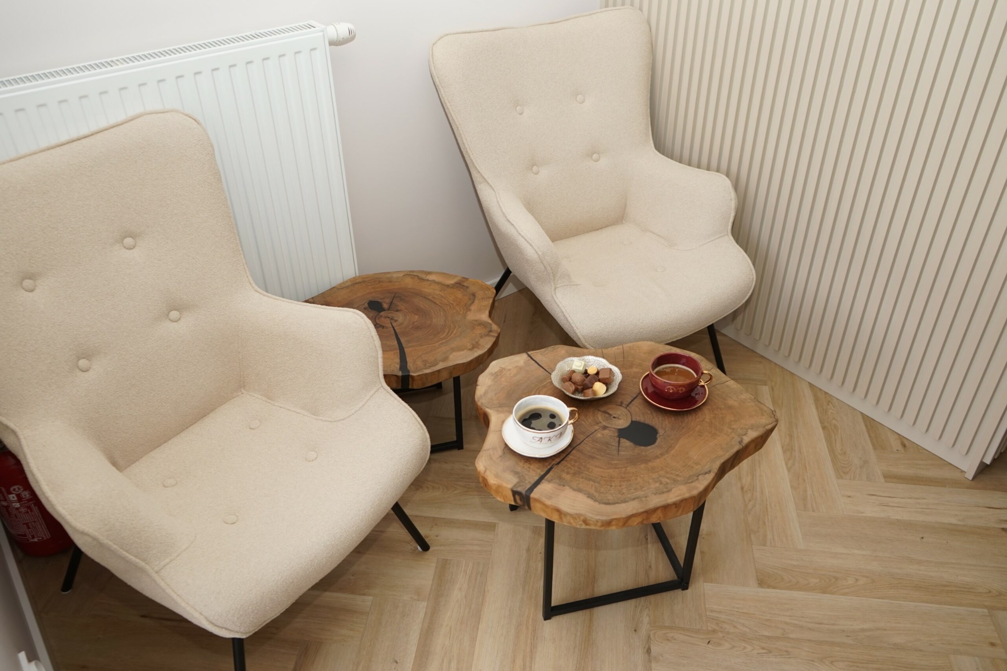 Dwa beżowe fotele klubowe z tapicerką boucle i okrągłe stoliki kawowe z plastra drewna z żywicą epoksydową, na podłodze ułożonej w jodełkę.