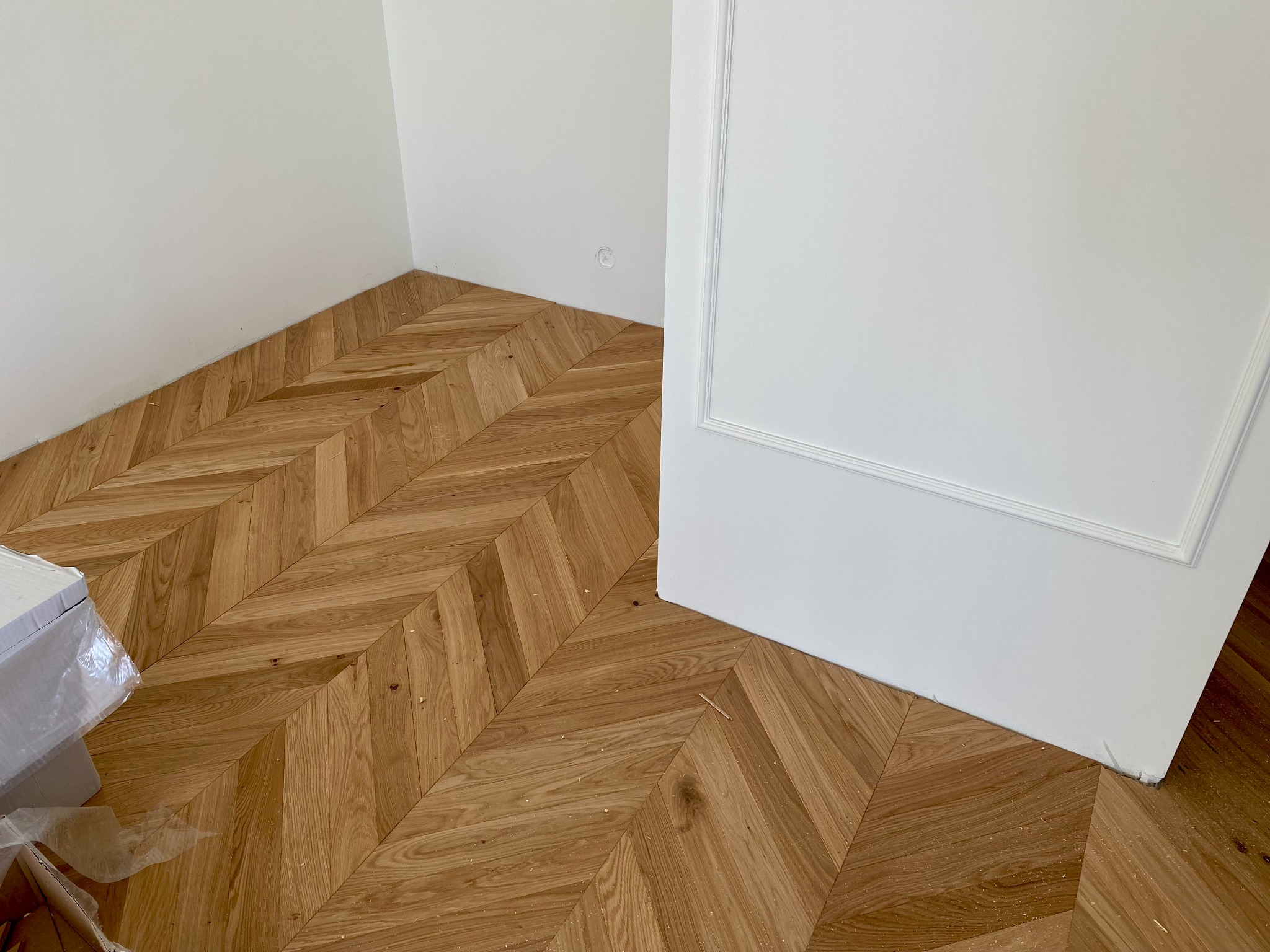Ułożony parkiet w jodełkę francuską, widoczne białe ściany i fragment białych drzwi z listwą dekoracyjną, karton z panelami w rogu.