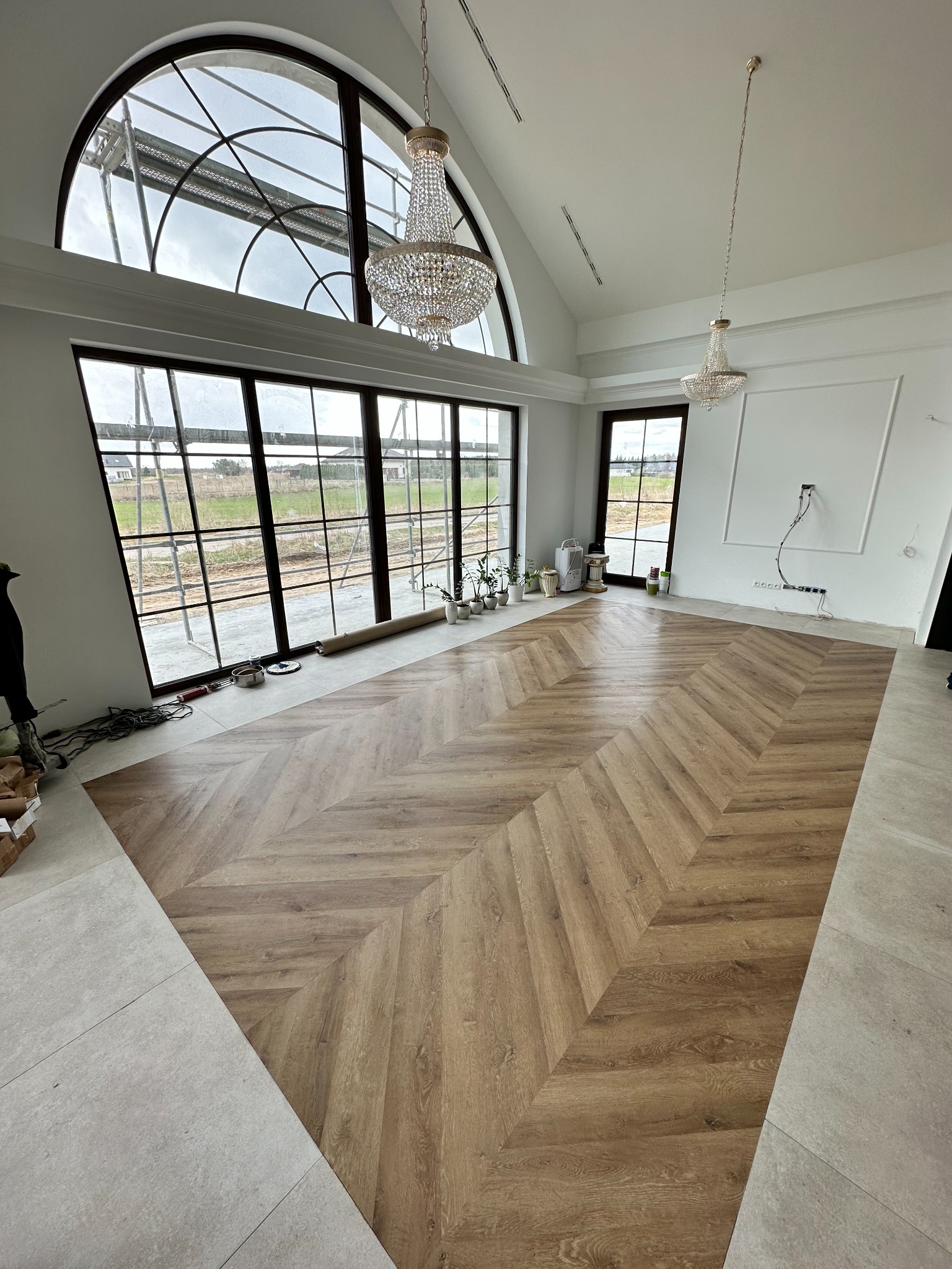Ukończona podłoga ułożona w jodełkę francuską w przestronnym salonie z dużymi oknami i żyrandolami, widoczne narzędzia i resztki materiałów