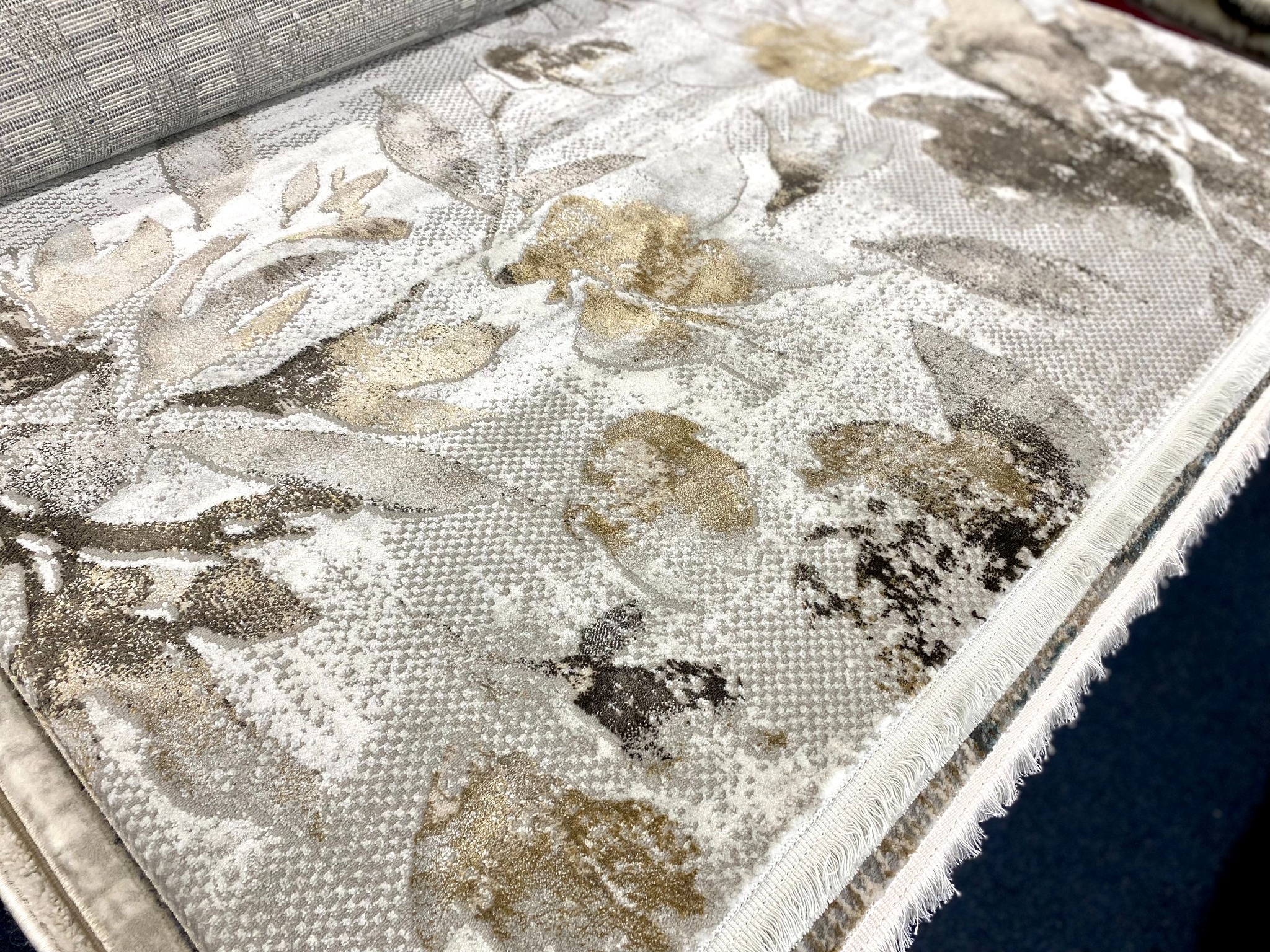Próbka dywanu z kwiatowym wzorem w odcieniach szarości, beżu i brązu, widoczna struktura i frędzle na krawędzi