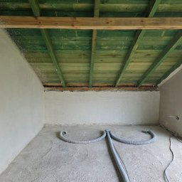 Majanek - Pokój w trakcie remontu: surowe ściany po szpachlowaniu, zielone drewniane belki stropowe, szare rury wentylacyjne na podłodze. Widok na okno.