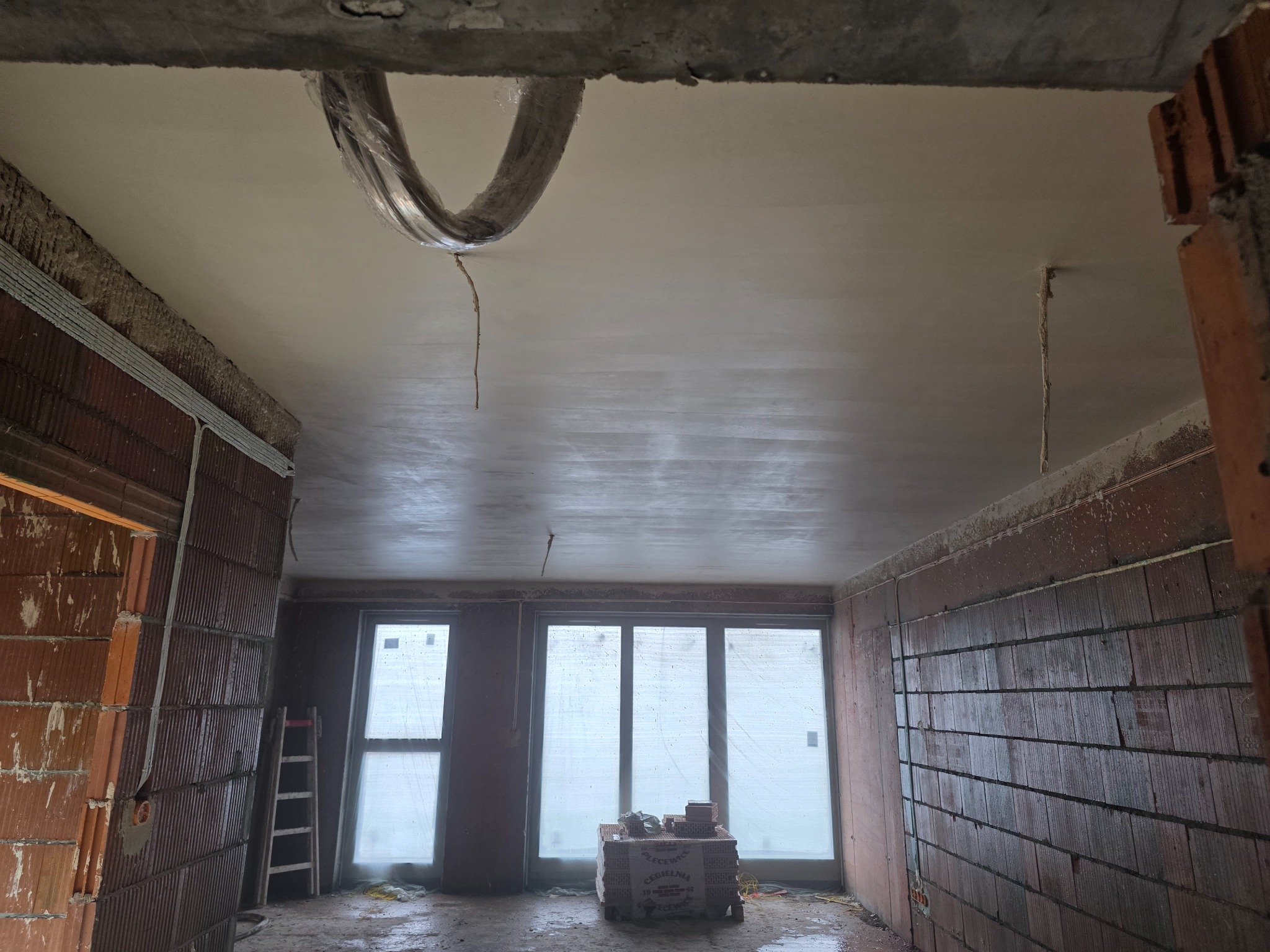 Wnętrze w trakcie prac wykończeniowych: sufit po gładziowaniu, ściany z cegły, okna zabezpieczone folią. Widoczne materiały budowlane i drabina.
