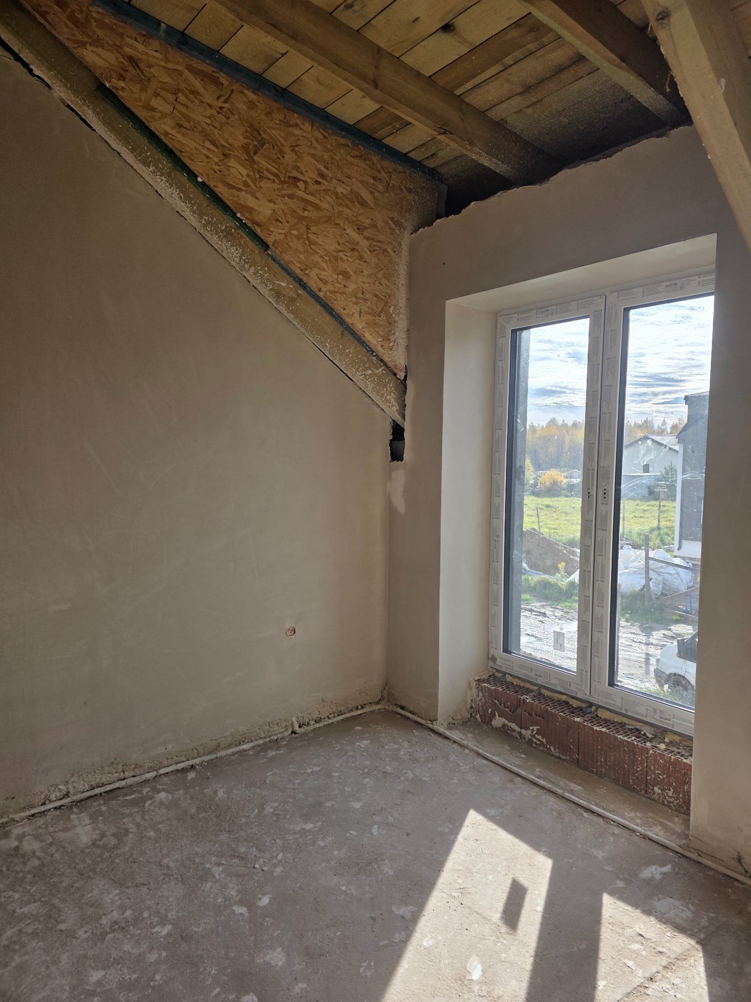 Pokój na poddaszu w trakcie szpachlowania ścian, widoczne drewniane elementy konstrukcyjne dachu i okno z widokiem na zewnątrz. Ściany w szarym kolorze, surowy beton na podłodze.