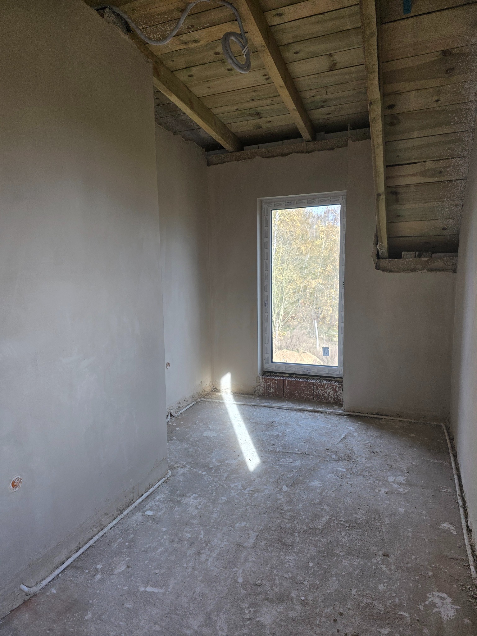Pokój po szpachlowaniu ścian, widoczne gładkie powierzchnie, surowy beton na podłodze, drewniany strop i okno z widokiem na drzewa. Promień światła na podłodze.