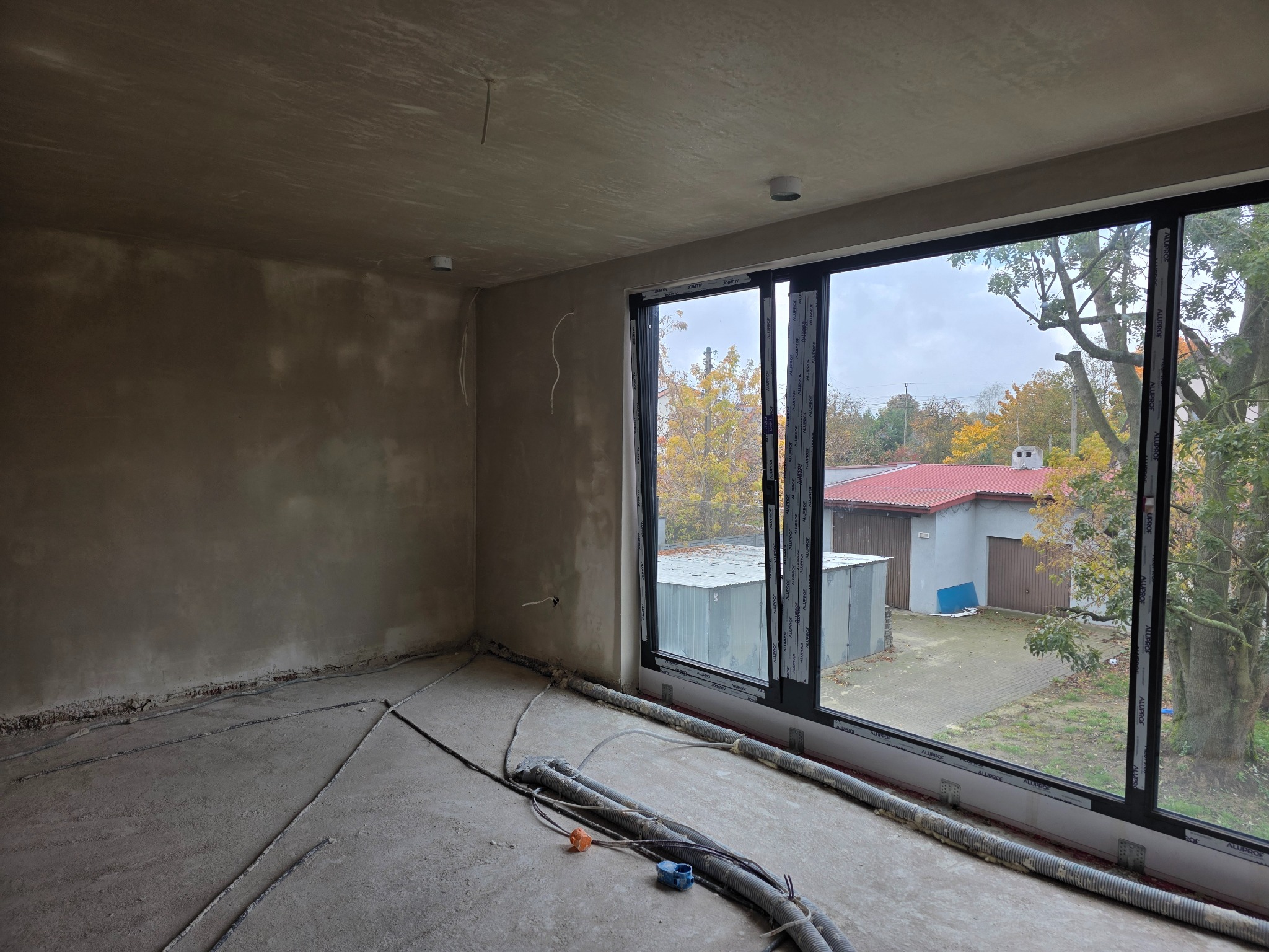 Surowe wnętrze z dużym oknem, ściany w szpachli, podłoga z instalacją ogrzewania podłogowego. Widok na zewnątrz z drzewami i budynkiem gospodarczym.
